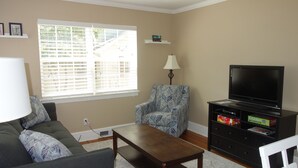 A living room with a sofa, armchair, coffee table, TV stand with a flat-screen TV, and a window with blinds. - The Better Half Duplex on 7th (Charlotte)