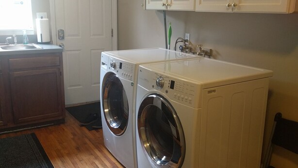 A laundry room with two front-loading washing machines, wooden cabinets, a sink, and a window. - The Better Half Duplex on 7th (Charlotte)