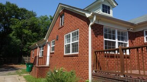 A red brick house with a white window trim, a wooden front porch, and a green trash bin. - The Better Half Duplex on 7th (Charlotte)