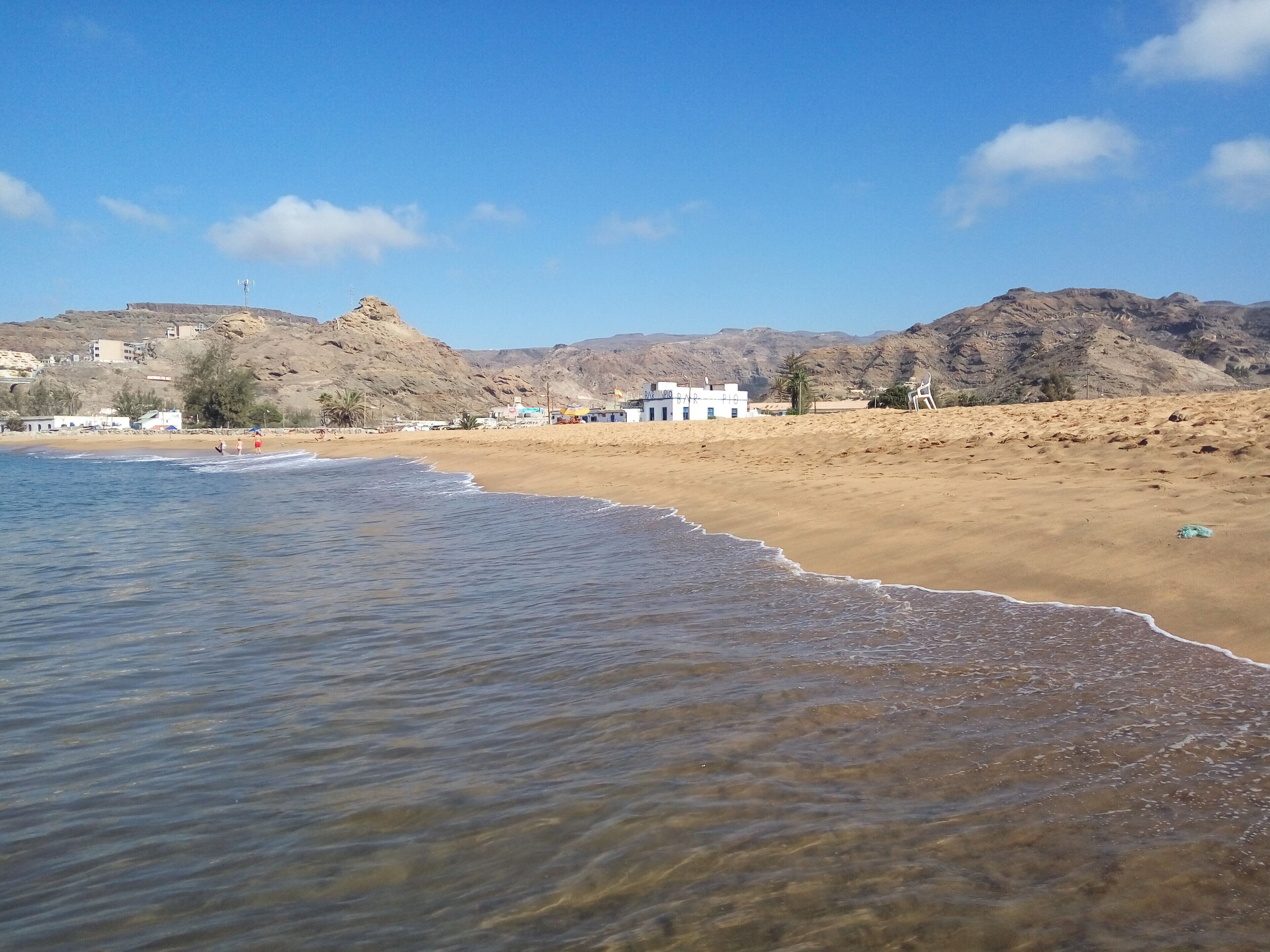 A beach with clear water, sandy shore, and buildings in the background.