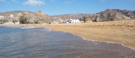 A beach with clear water, sandy shore, and buildings in the background.
