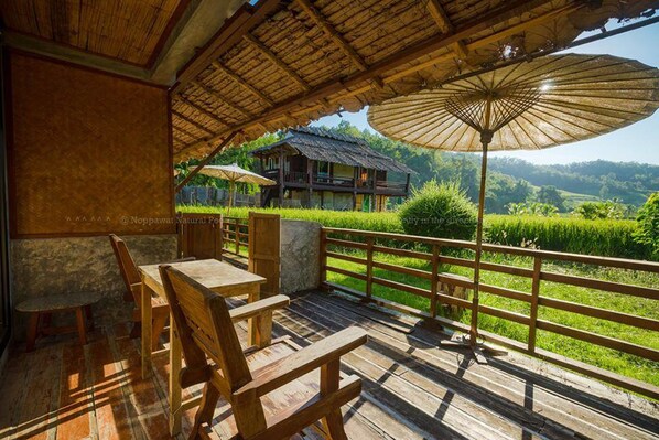 A wooden deck with outdoor seating, a thatched roof, and a view of a house and lush greenery.
