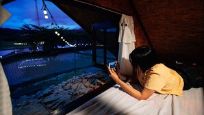 A person lying on a bed in a tent-like room, looking out at a body of water with lights in the distance.