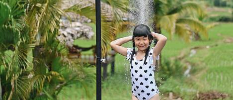 Un niño bajo una ducha en un jardín con palmeras y un paisaje montañoso.