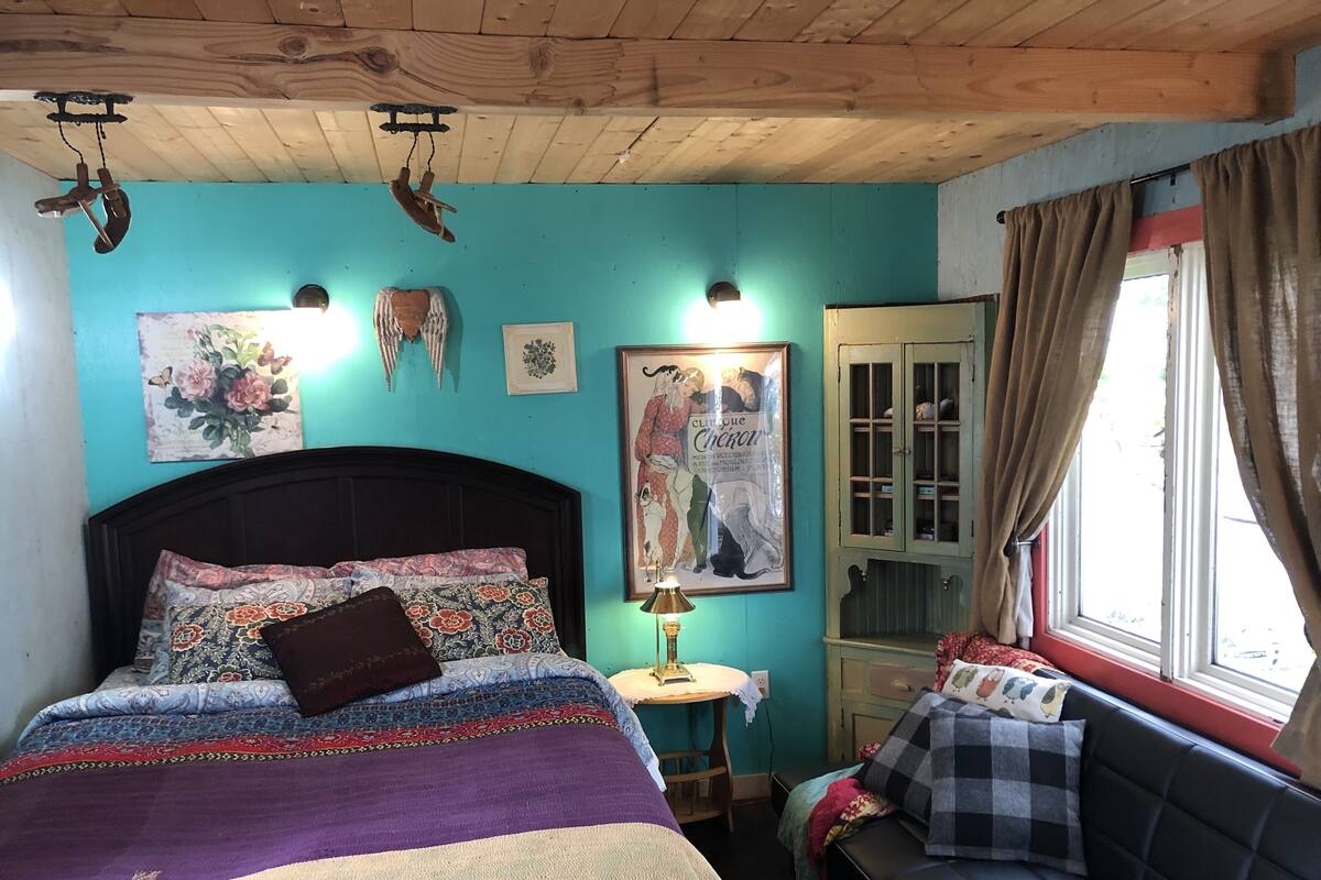 A bedroom with a wooden ceiling, a bed with patterned bedding, a small table with a lamp, a window with curtains, and a wall with framed artwork.