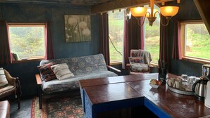 A cozy room with a wooden ceiling, a sofa with floral upholstery, a dining table with a tray of cups and a kettle, and a view of greenery through large windows.