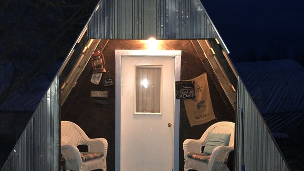 A small, triangular metal structure with a white door, two wicker chairs, and a welcome sign.