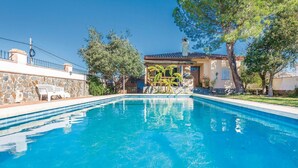 Una piscina con un muro de piedra y una banca, rodeada de árboles y un edificio con techo de tejas.