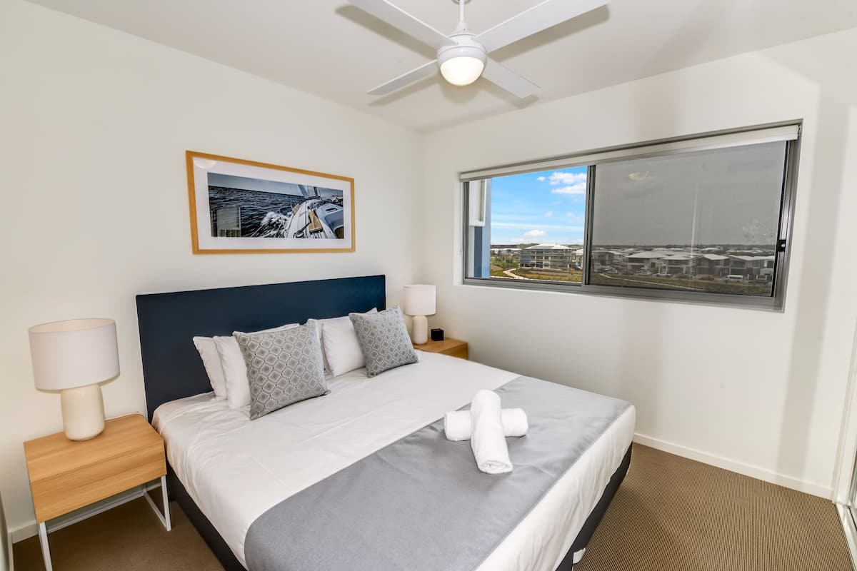 Une chambre d’hôtel avec un lit, un ventilateur de plafond, une fenêtre donnant sur un beau paysage, un tableau encadré au mur et une table de chevet avec une lampe.