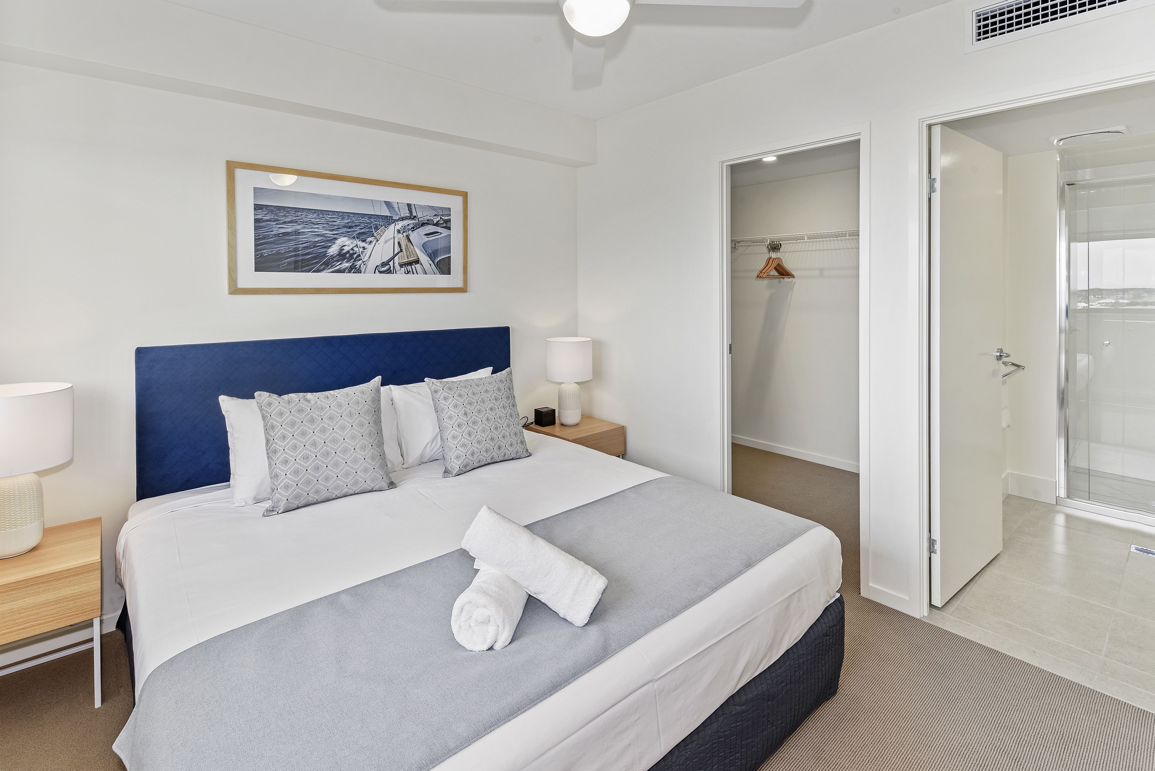 Een netjes opgemaakt bed met een blauw hoofdeinde, een ingelijst kunstwerk aan de muur, en een badkamer met een douchecabine van glas.