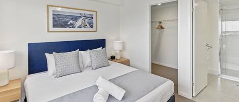 Una cama bien hecha con cabecera azul, un cuadro en la pared y un baño con ducha de vidrio.
