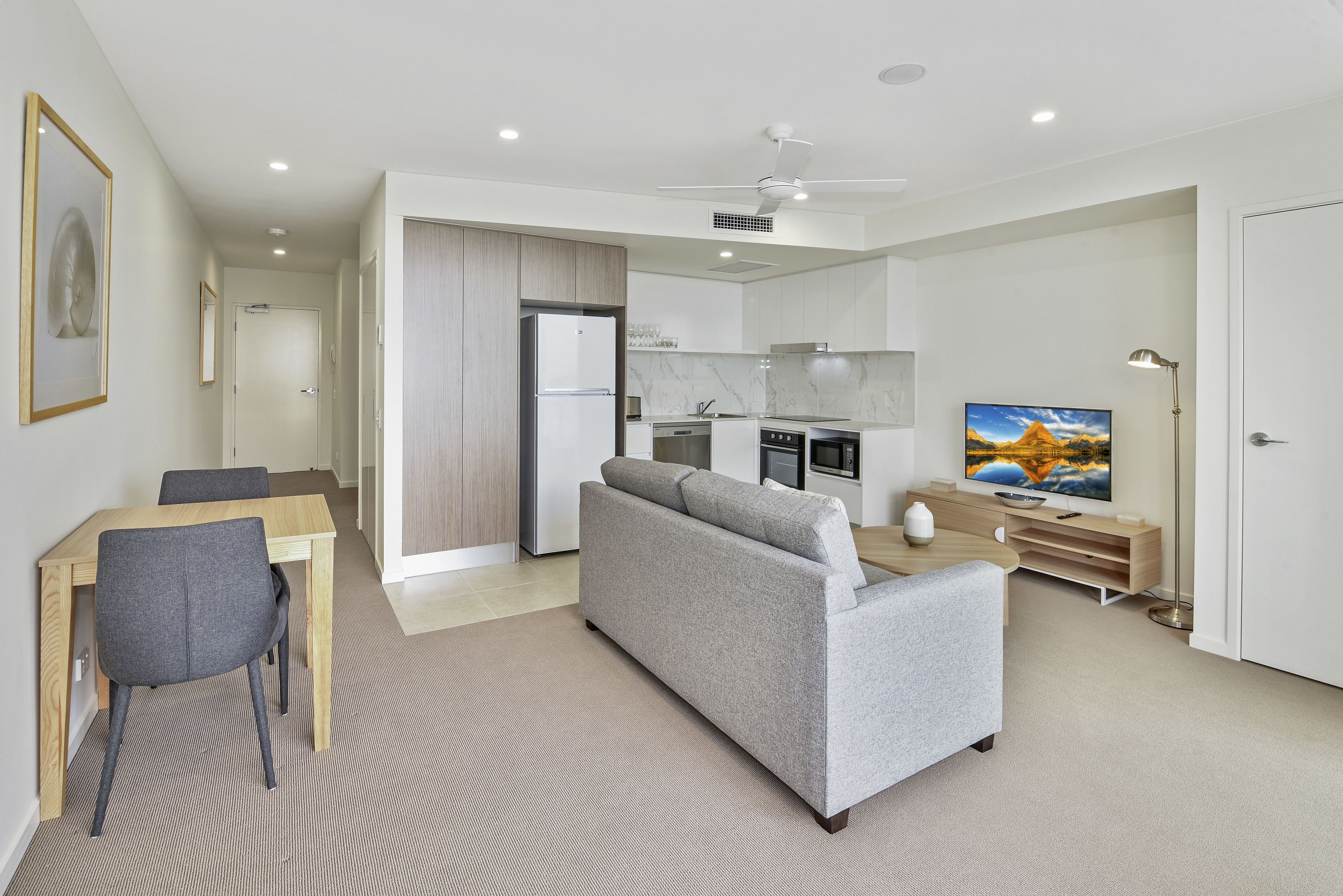 A modern living room with a grey sofa, a wooden dining table with two chairs, a kitchen area with a fridge, stove, and microwave, and a flat-screen TV on a stand.