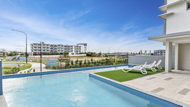 A modern swimming pool with a clear blue water, surrounded by a tiled deck and a grassy area with lounge chairs. In the background, there is a multi-story building with balconies.