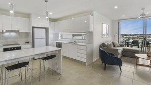 A modern kitchen with white cabinets, a central island with bar stools, a built-in dishwasher, and a large window offering a city view.