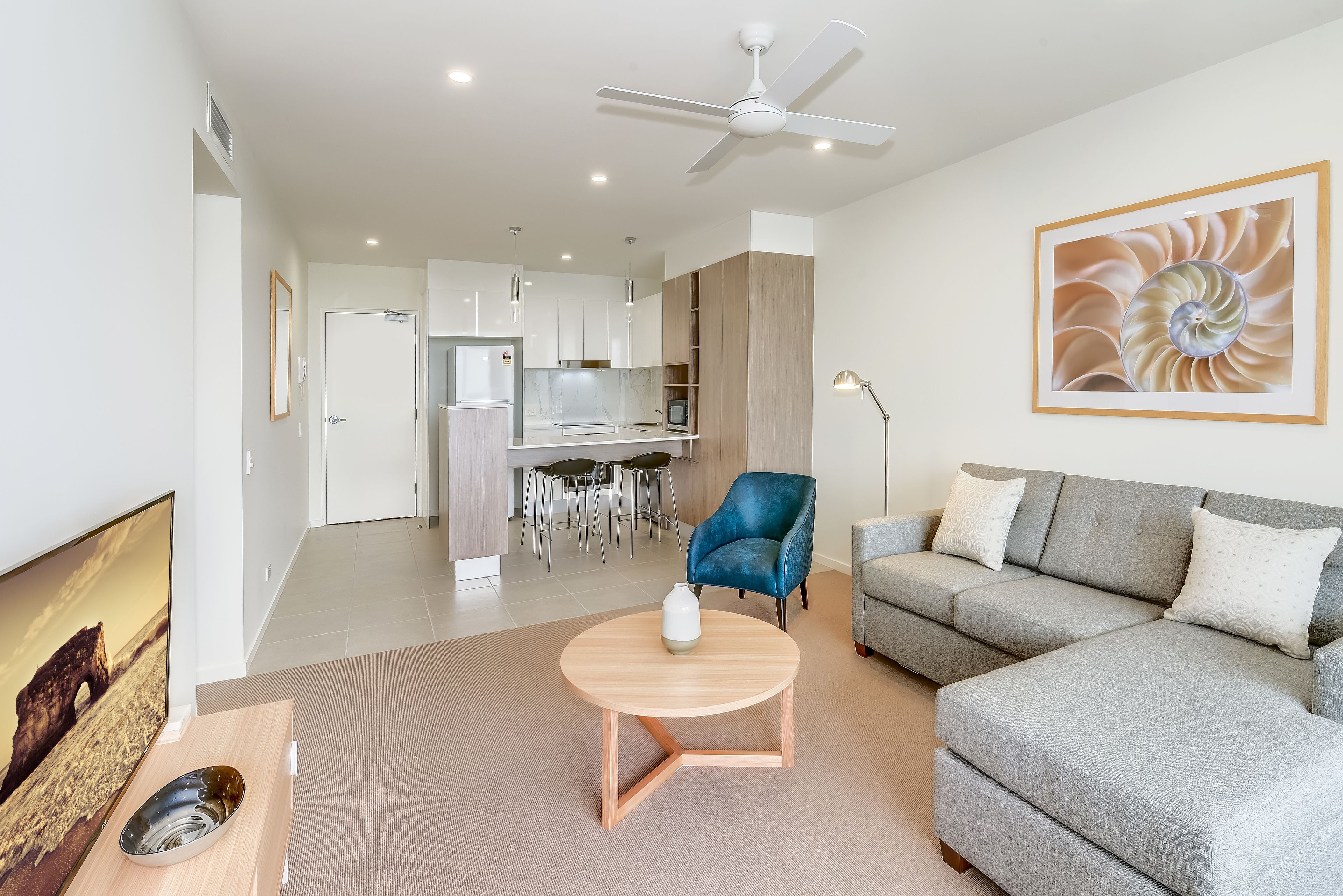 A modern living room with a grey sofa, a round wooden coffee table, and a blue armchair. A kitchen is visible in the background with white cabinets and a dining area.