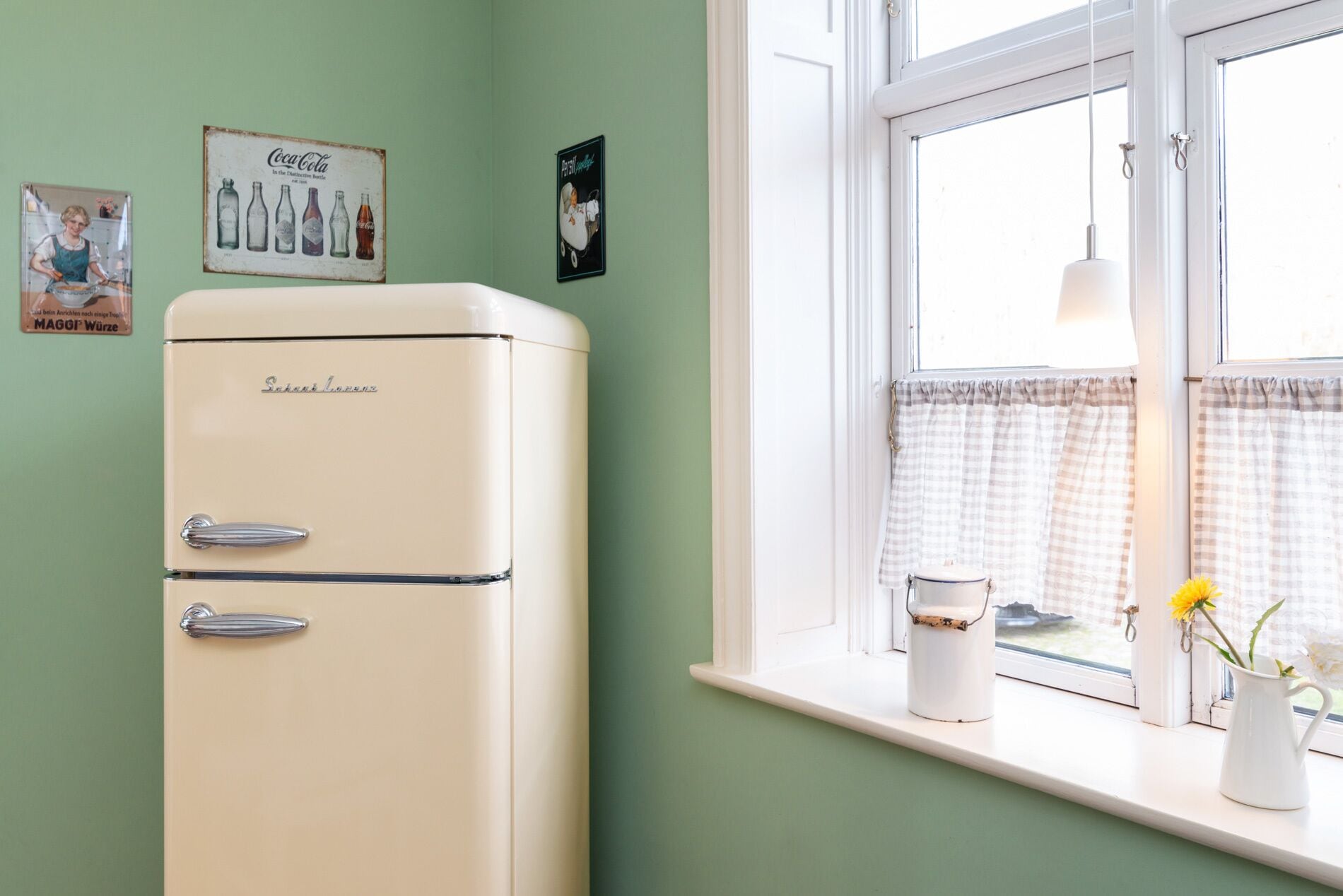 A vintage refrigerator in a room with a window, a checkered curtain, and a vase with flowers on the windowsill.