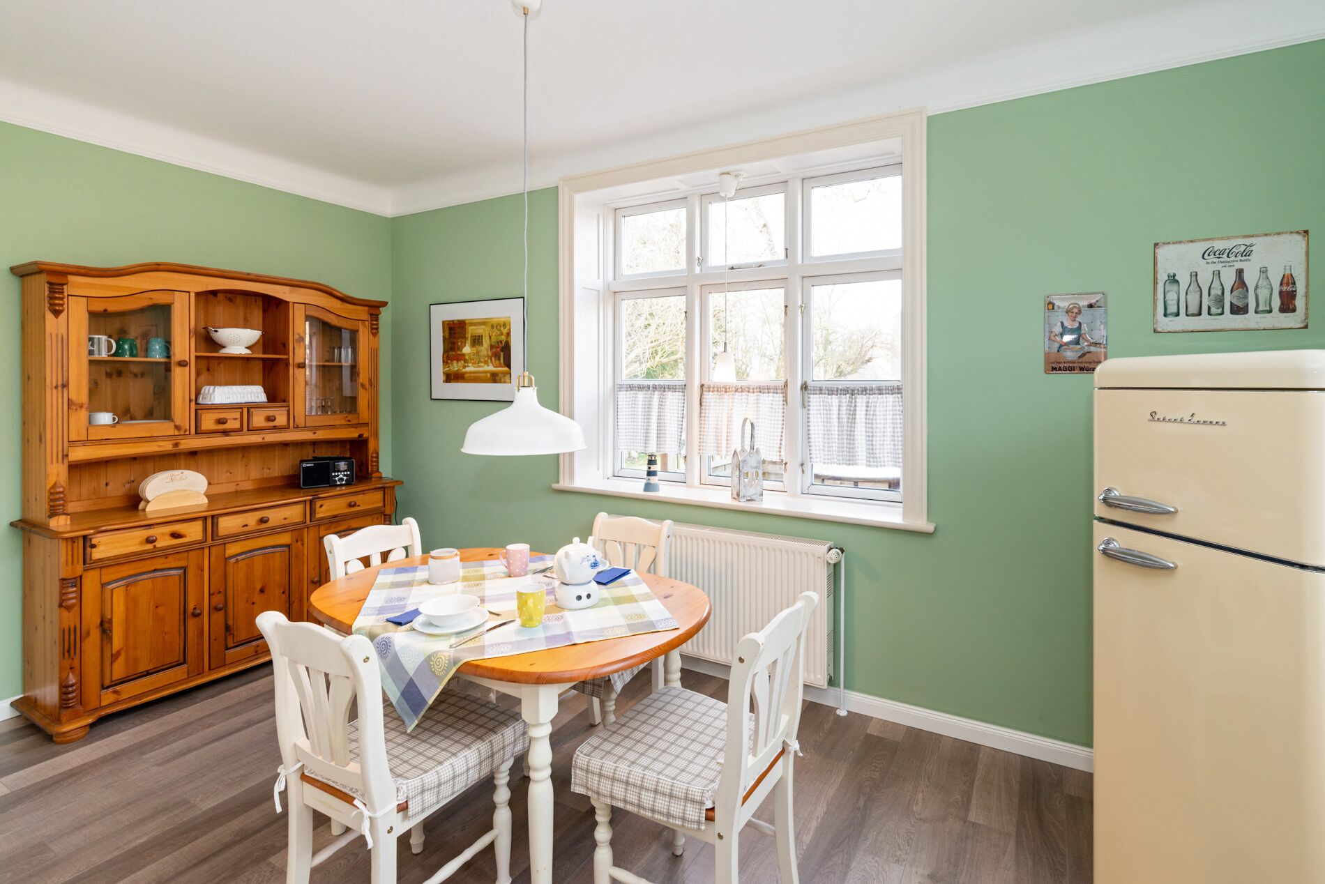 A dining area with a wooden cabinet, a table with chairs, a vintage refrigerator, and a window with curtains.