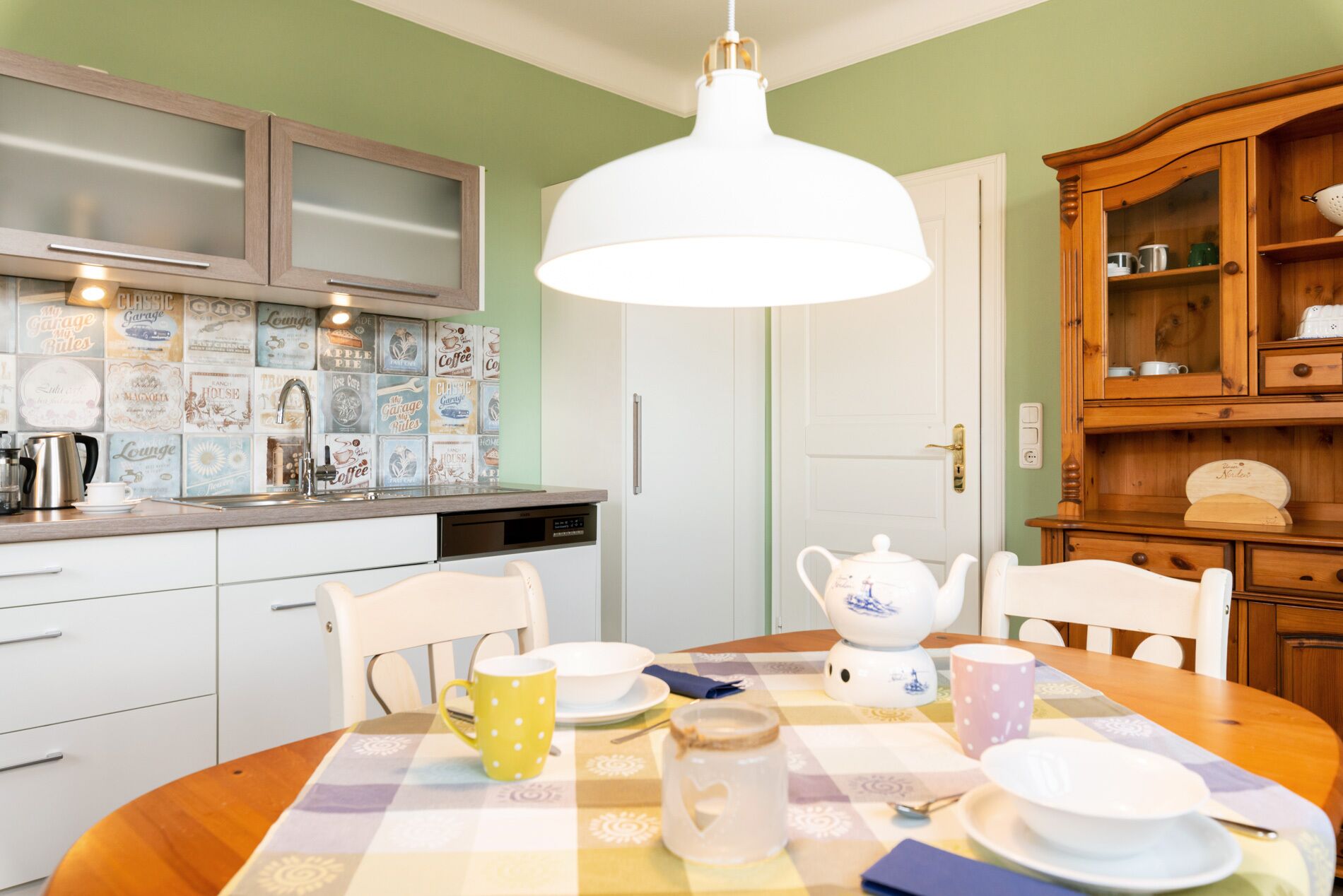 Een keuken met een wit spoelgedeelte, een koffieapparaat en een eettafel gedekt met borden, kopjes en een theepot.