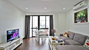 A living room with a TV, a sofa, a coffee table with books and a vase, a dining table with chairs, and a large window with curtains.