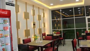 A dining area with tables and chairs, a Coca-Cola refrigerator, and a large window with a view. - Orient Hotel (Mandalay)