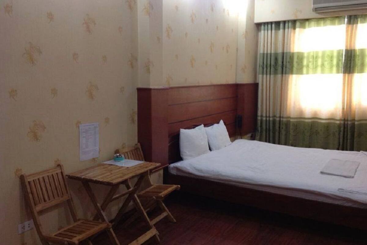 A hotel room with a single bed, a wooden desk, two chairs, and a window with curtains.