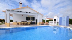 Una piscina con un patio cubierto, un edificio blanco con techo de tejas y un borde de piscina con azulejos azules.