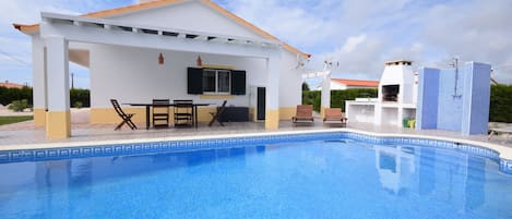 A swimming pool with a covered patio area, a white building with a tiled roof, and a blue-tiled pool edge.
