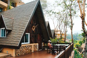 A wooden cabin with a sloped roof and stone wall, a deck with a table and chairs, and a view of the surrounding forest. - Suansaiyok Resort (Sai Yok)