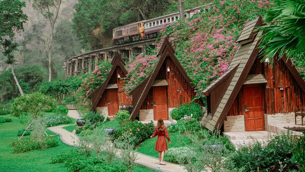 A row of wooden cabins with a garden path leading to them, surrounded by lush greenery and a train in the background. - Suansaiyok Resort (Sai Yok)