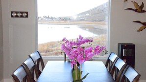Une table à manger avec des chaises, un vase rempli de fleurs violettes et, par la fenêtre, une vue sur un lac et des collines.