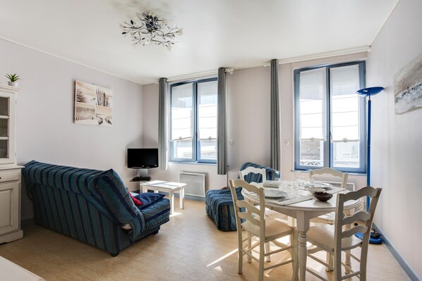 Una habitación bien iluminada con una mesa de comedor dispuesta para dos personas, un sofá, un televisor y ventanas grandes con cortinas.