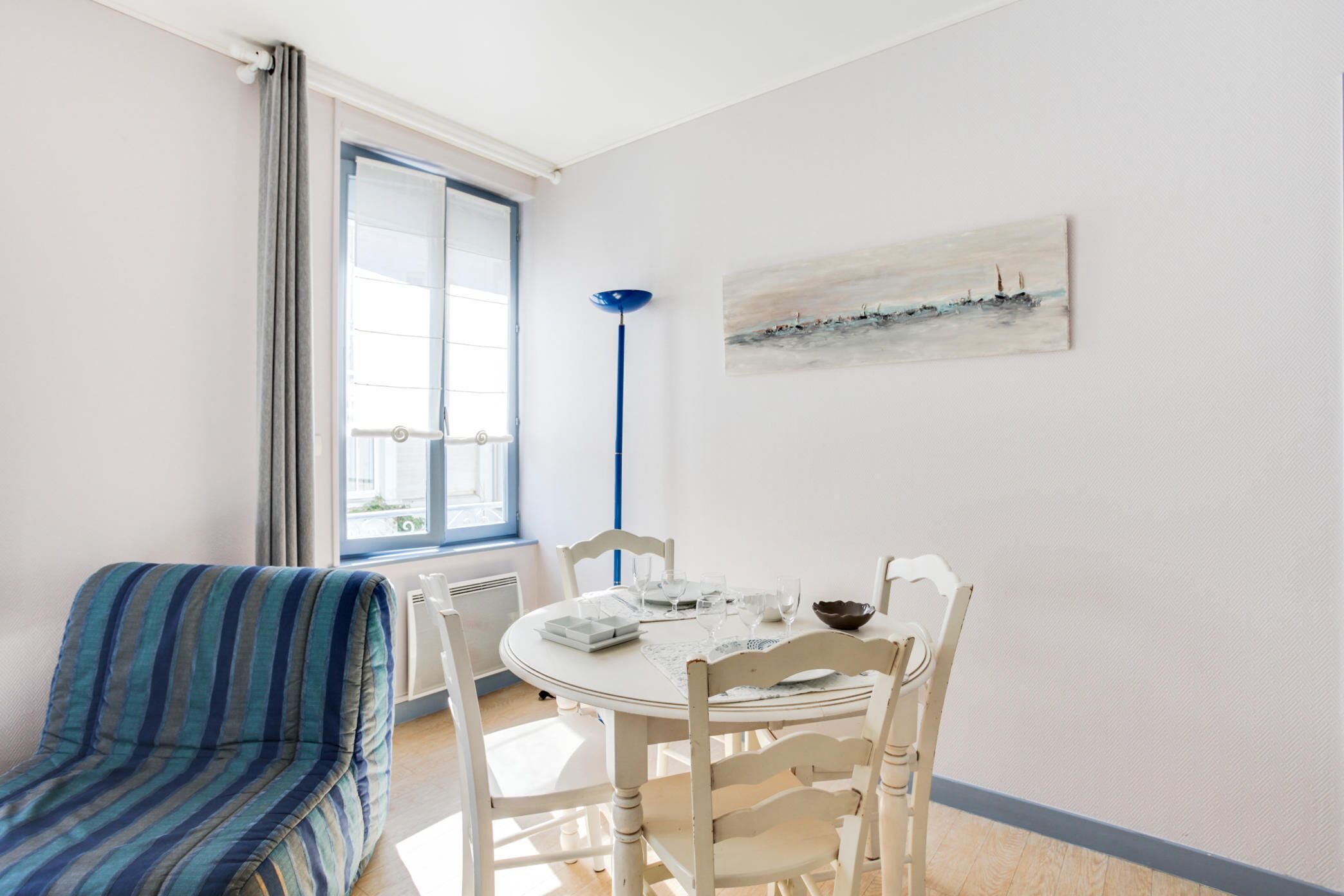 Een eetgedeelte met een ronde tafel, witte stoelen, een gestreept sofa, een schilderij aan de muur en een raam met gordijnen.