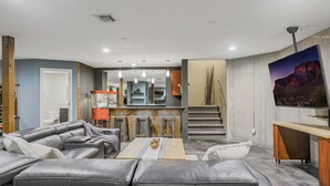 A modern living room with a leather sofa, a coffee table, and a TV. There is a kitchen area with a counter and cabinets, and a staircase leading to another level.