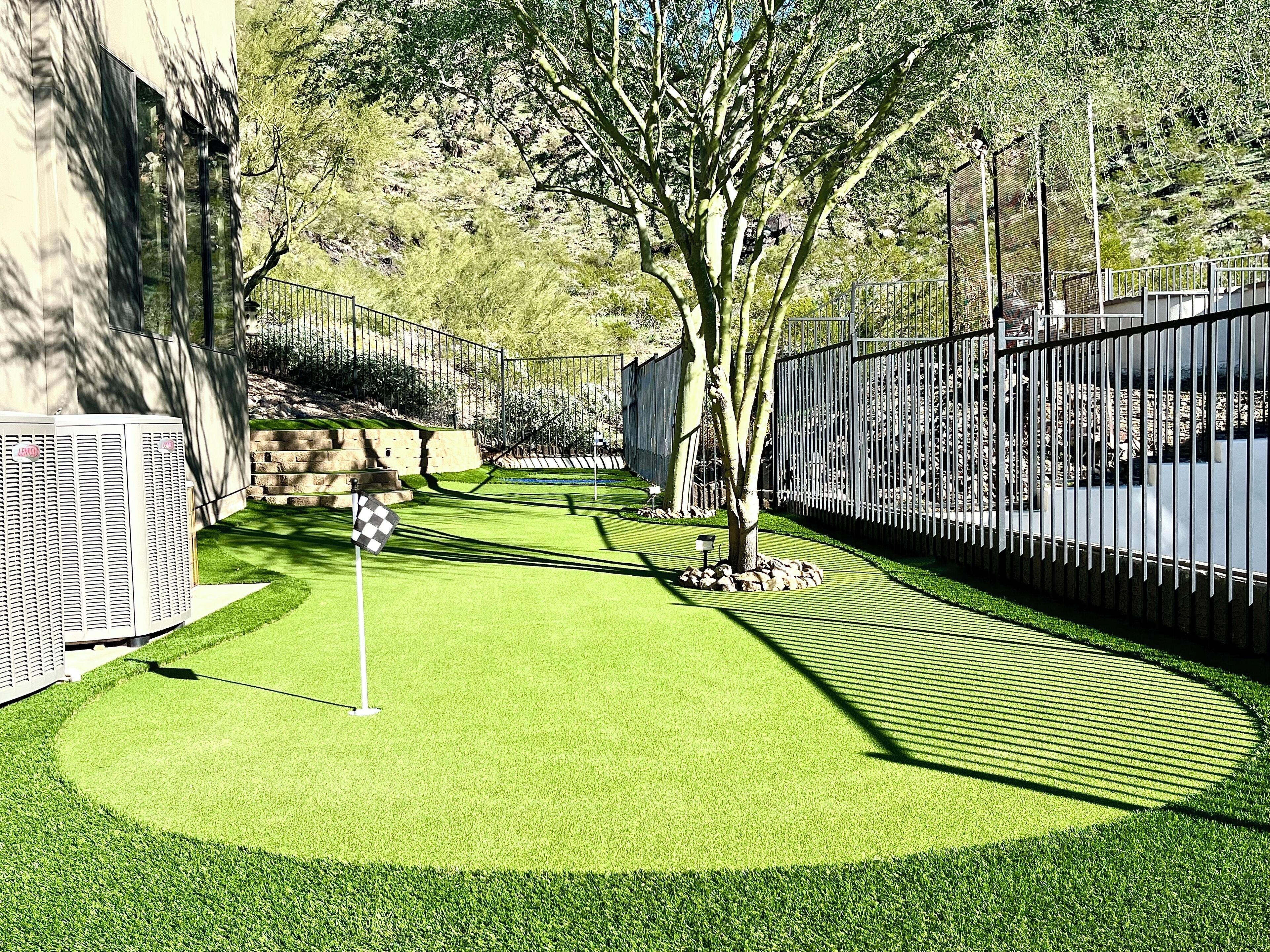 Un campo de golf de césped artificial bien cuidado, con un área de práctica de putting, rodeado por una valla de metal y árboles.