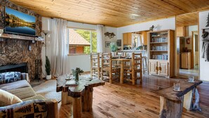 A cozy cabin interior with a stone fireplace, wooden dining table, and a kitchen area.