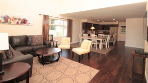 Ein Wohnzimmer mit Sofa, Couchtisch und Sesseln, ein Essbereich mit Tisch und Stühlen sowie eine Küche im Hintergrund.