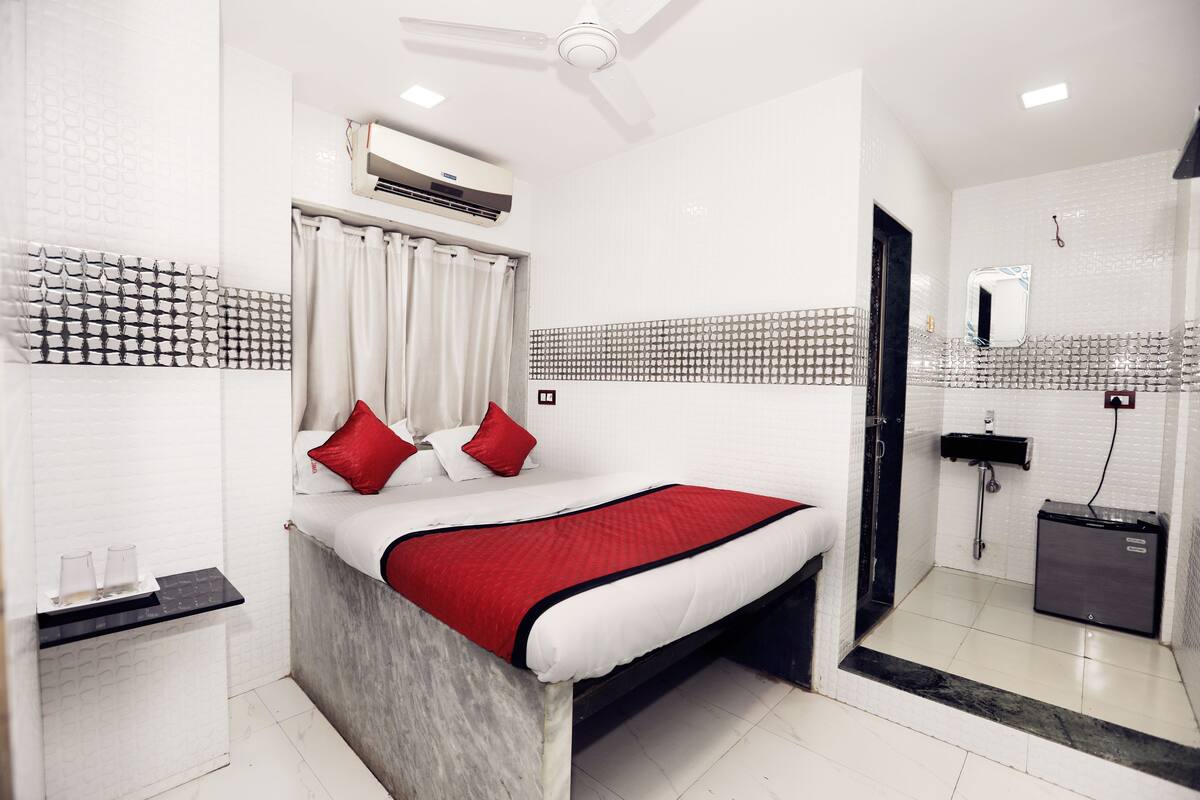 A modern hotel room with a large bed, a bedside table with a lamp, a built-in air conditioner, a shower area, and a small storage cabinet.