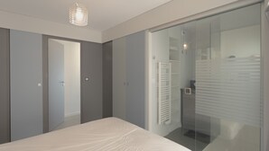 A modern bedroom with a large bed, a sliding glass door leading to a bathroom, and a wardrobe with a mirror. - Seaside Apartment with Balcony, Close to Shops (Les Sables-d'Olonne)