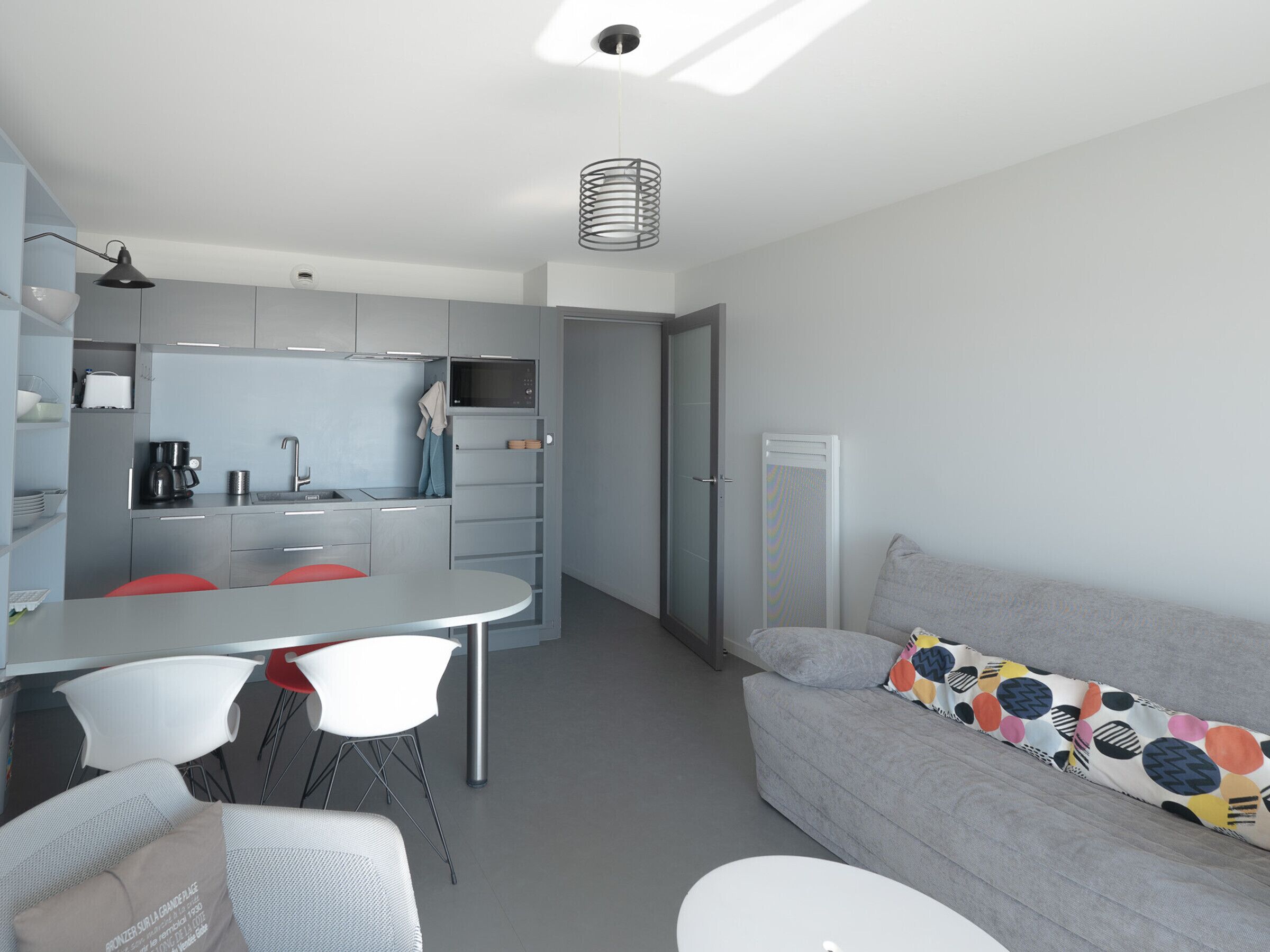 Un espacio habitable pequeño con kitchenette, una zona de comedor con sillas, un sofá con un cojín colorido y un aplique de techo.