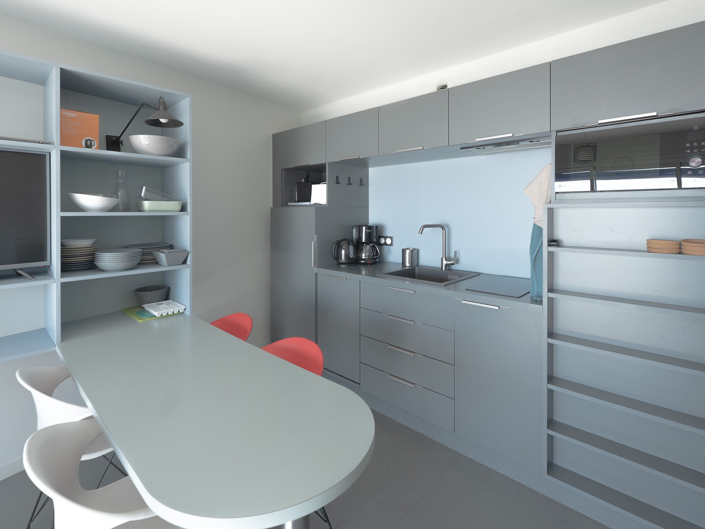 A modern kitchen with grey cabinets, a white countertop, and a built-in oven. There are two red chairs and a white table.