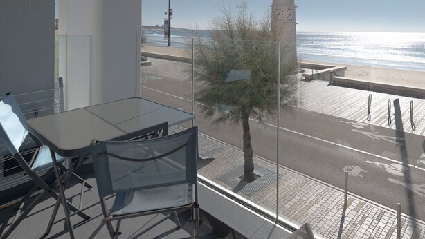 A balcony with a glass table and chairs, overlooking a beach with a clock tower, a street, and a pedestrian path. - Seaside Apartment with Balcony, Close to Shops (Les Sables-d'Olonne)