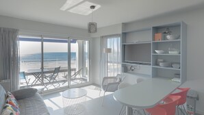 A modern living room with a large window overlooking the beach, a dining area with a white table and red chairs, and a kitchen with open shelving. - Seaside Apartment with Balcony, Close to Shops (Les Sables-d'Olonne)