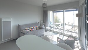 A modern hotel room with a grey sofa, a small round table, and a balcony with outdoor seating. - Seaside Apartment with Balcony, Close to Shops (Les Sables-d'Olonne)