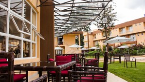 An outdoor seating area with wicker furniture and red cushions, a glass ceiling structure, and a view of a building with balconies. - Elysian Resort (Nairobi)