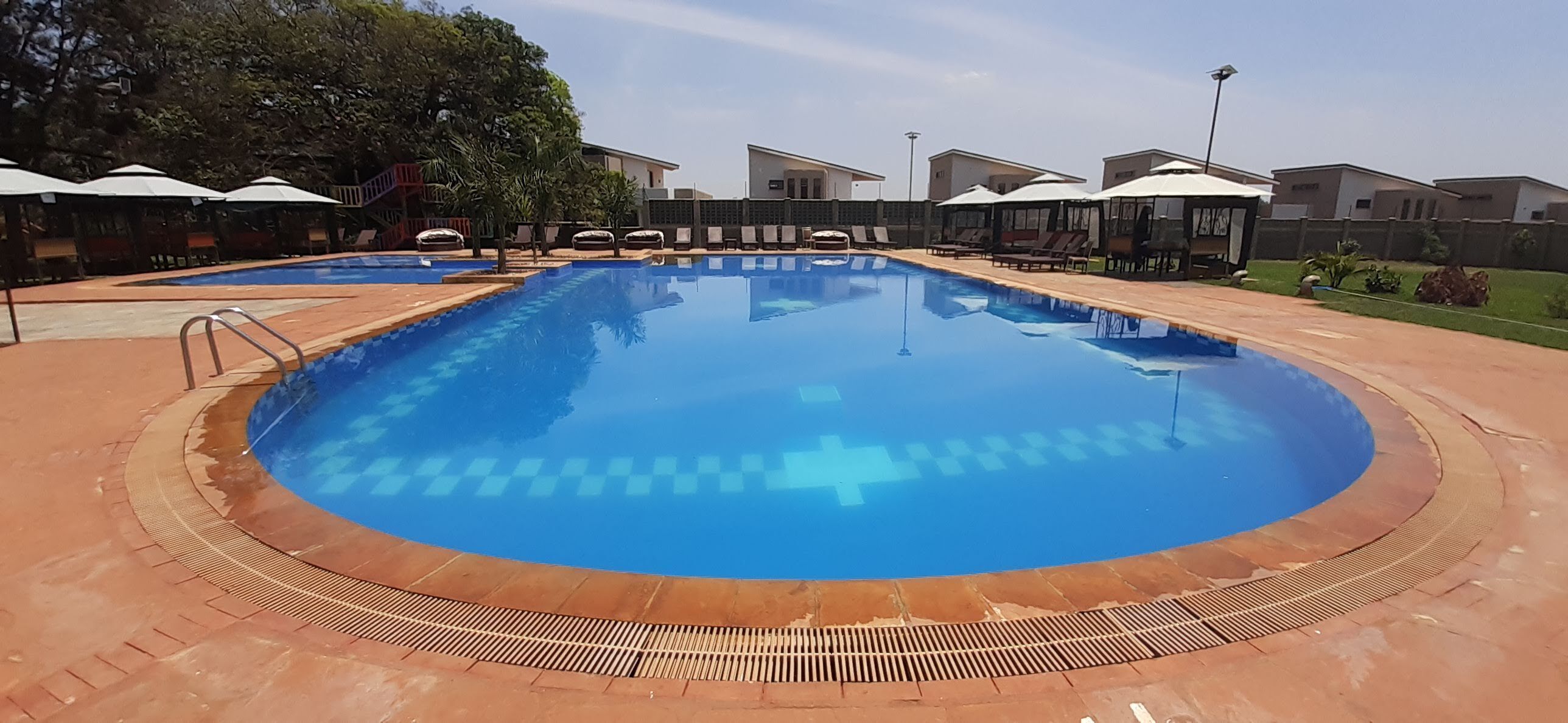 Una piscina con cubierta de baldosas, rodeada de sillas de descanso y sombrillas, con edificios al fondo.