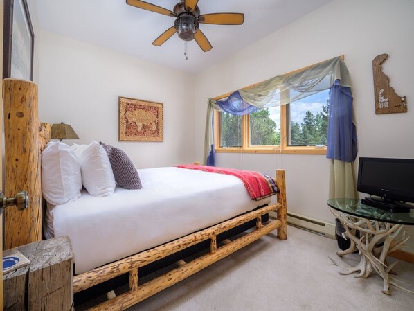 A bedroom with a wooden bed frame, a ceiling fan, a window with curtains, a television on a glass table, and a wall decoration. - RMR: Strawberry 3921 Condo in The Aspens/Wilson,WY (Wilson)