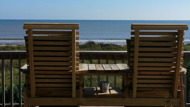 Dos sillones de madera mirando una playa con una terraza de madera y una pequeña mesa con un cubo y una taza.
