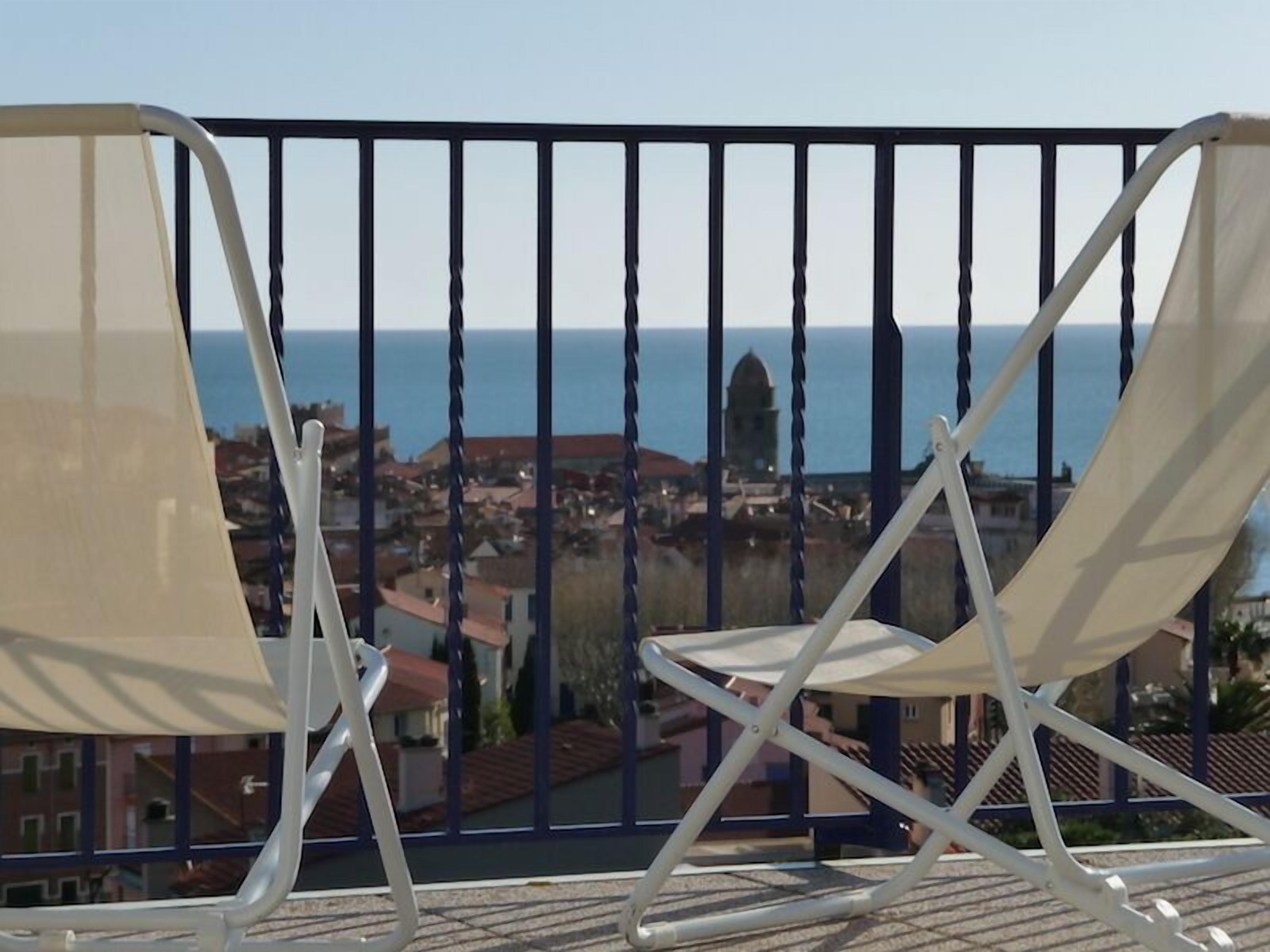 A balcony with two white folding chairs, a metal railing, and a view of the sea and buildings.