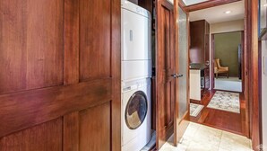 Een wasserette met een wasmachine en droger, houten kasten en een deur naar een woonruimte met een stoel en vloerkleed.