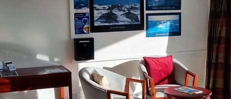 A hotel room with a desk, a chair, a small table, and a wall adorned with framed pictures of ocean scenes.